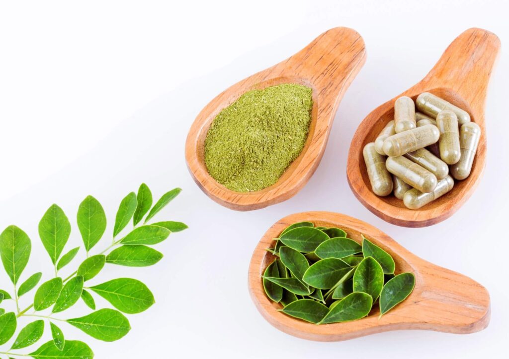 Moringa superfood in three forms – fresh leaves, powdered supplement, and capsules – displayed in wooden spoons, showcasing its versatility and health benefits.