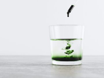 قطرات من مكمل الكلوروفيل السائل تُضاف إلى كوب ماء، مما يخلق نمطًا أخضر جذابًا – مشروب طبيعي لإزالة السموم وتعزيز الطاقة.